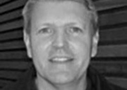 The image shows a grayscale portrait of a smiling person with short hair, standing in front of a striped background. The person wears a dark top.