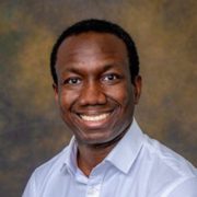This is a headshot of an adult person with a cheerful smile, wearing a light blue shirt, set against a mottled brown and yellow background.