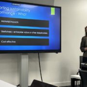 A person presents a slideshow titled "Reporting sustainability performance" in a corporate setting, discussing material impacts, usefulness, and cost-effectiveness.