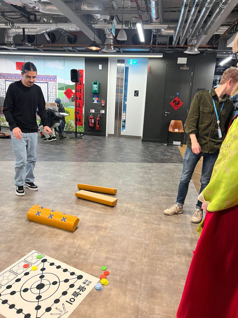 Two people are standing in a modern indoor space with exposed ceiling pipes. There's a board game on the floor and colorful stools nearby.