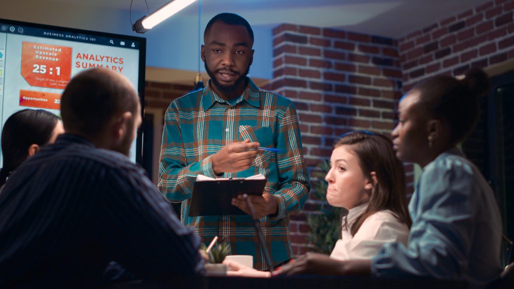 A person is presenting to colleagues using a digital screen with analytics data in a modern office setting with brick walls and indoor plants.