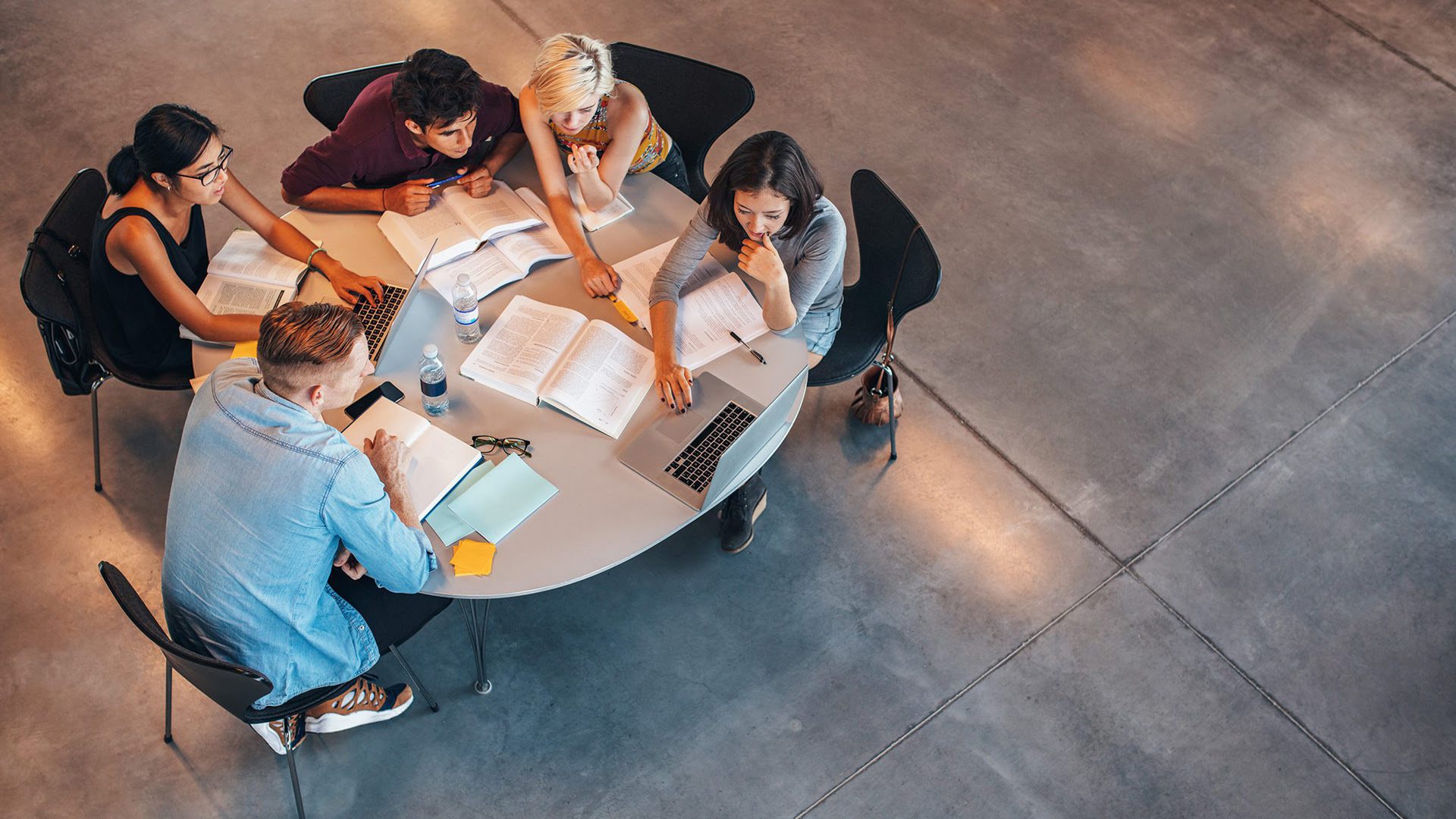 Five people gathered around a circular table are engaged in a study session with books, notes, and a laptop, in a well-lit modern space.