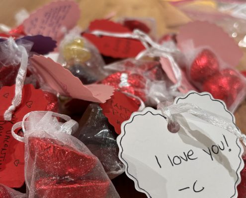 The image shows a collection of red heart-shaped chocolates with personalized notes attached, expressing love and affection, possibly for Valentine's Day.