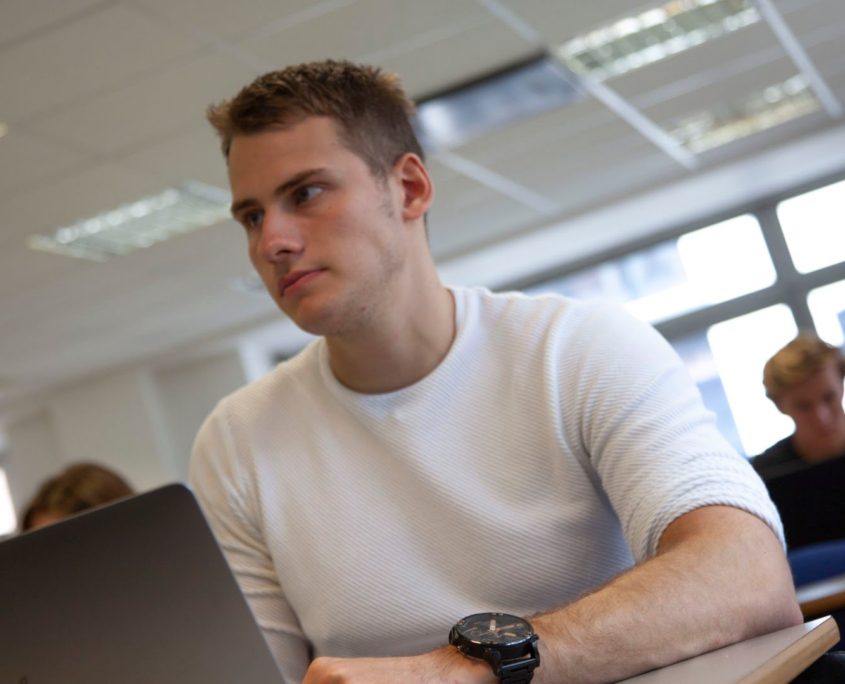 A focused person sits in a classroom with a laptop, wearing a white shirt and a watch, with other attendees in the background.