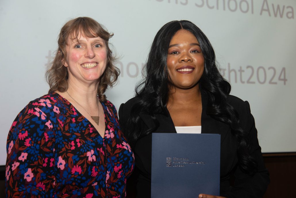 Two people are smiling in a room; one is holding a certificate. There's a projected text background, possibly from an awards ceremony.