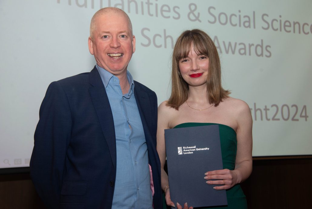 Two people are smiling in a room with a "Humanities & Social Sciences Awards" sign. One holds an award from Richmond American University London.