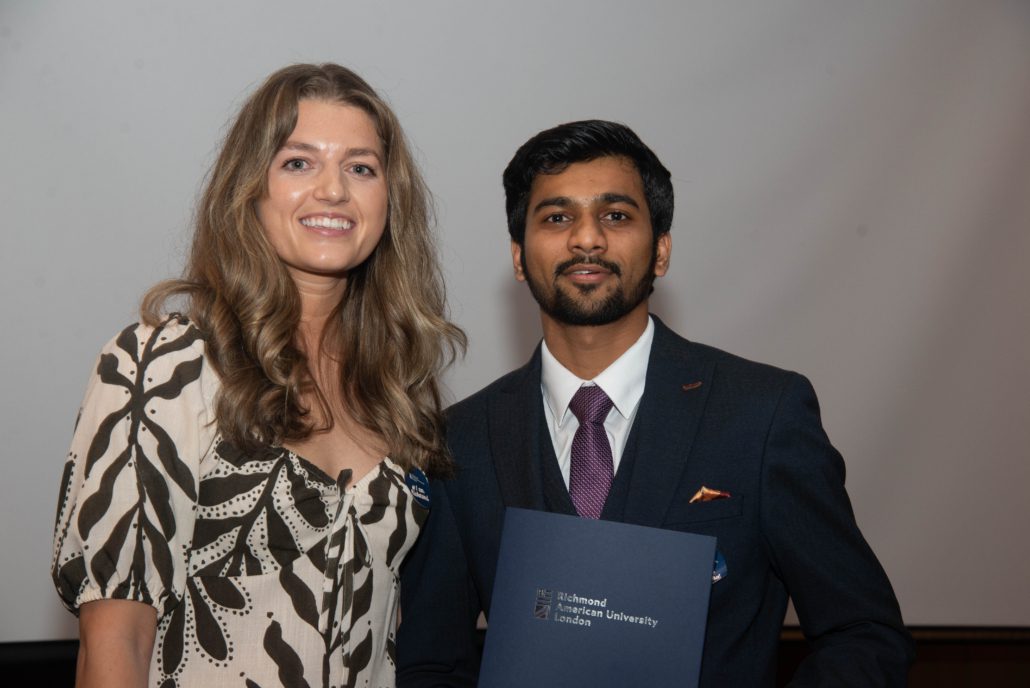 Two people are posing with a certificate from Richmond, The American University in London. One person is wearing a patterned blouse; the other, a suit.