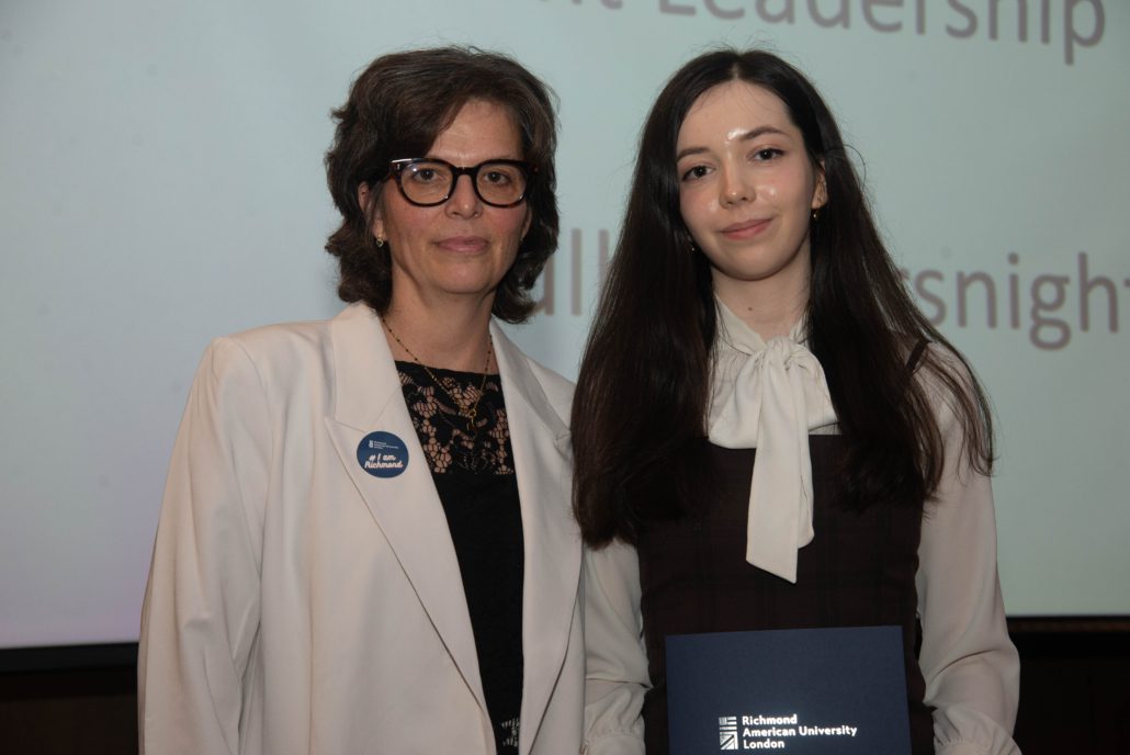 Two people are posing indoors, with one holding a “Richmond American University London” booklet. They're smiling in front of a presentation titled "Insights Leadership."