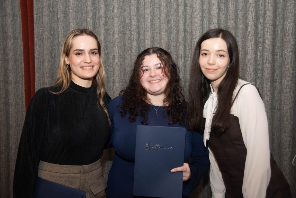 Three people are posing for a photo in front of a grey curtain; the middle person is holding a blue folder with embossed text. They are smiling.