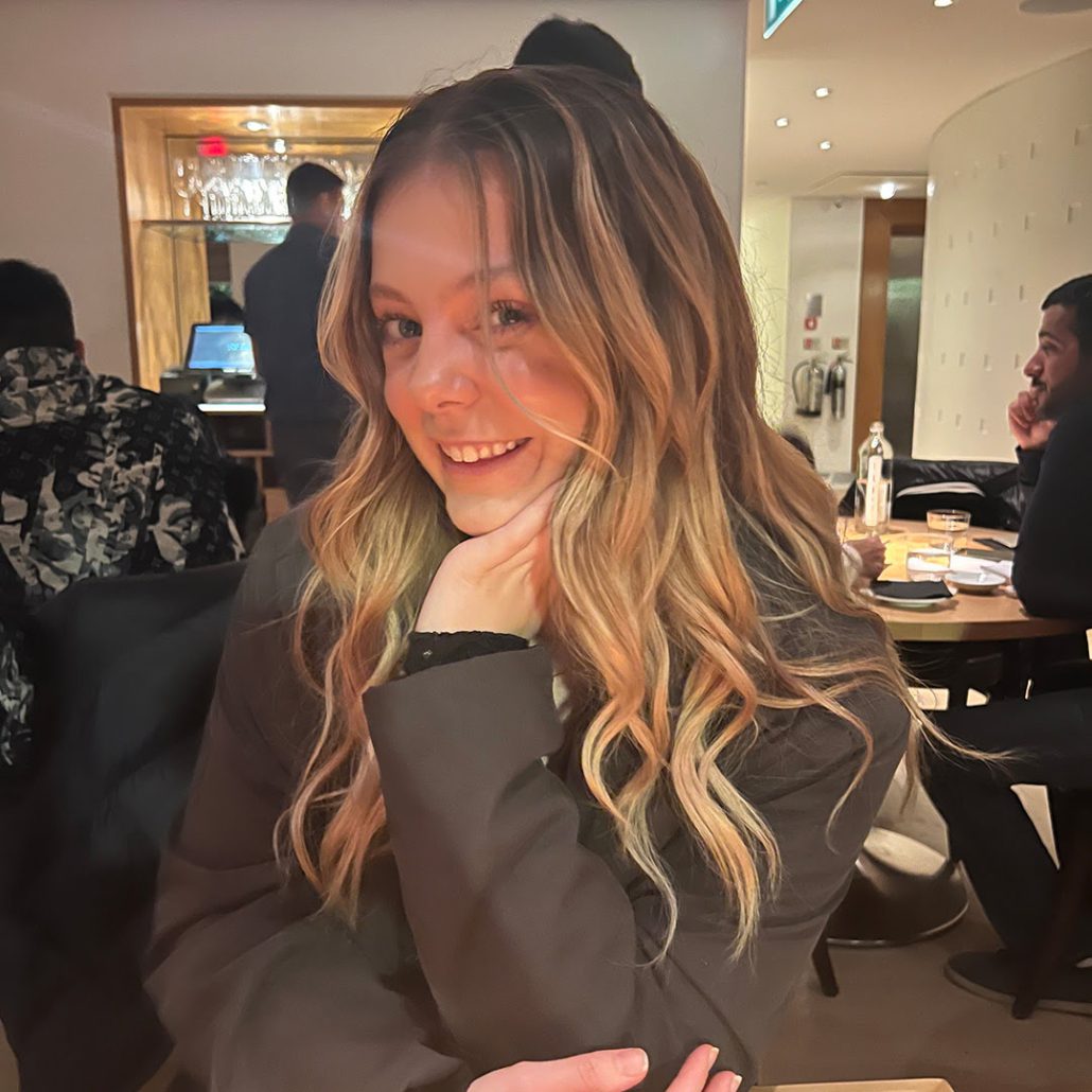A smiling person is posing with their chin resting on their hand at a restaurant, with warm lighting and other diners in the background.