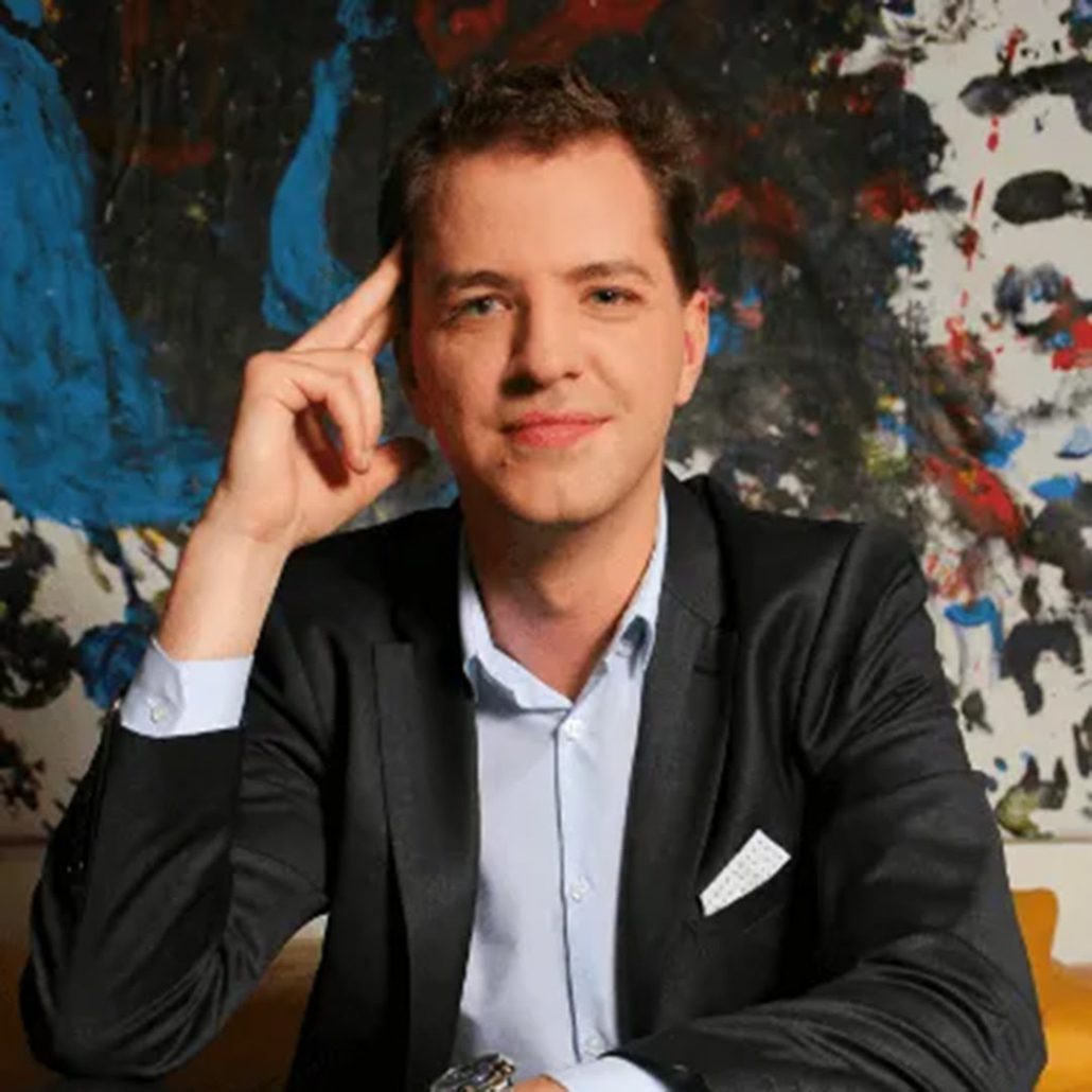 A person in a black blazer and blue shirt poses confidently, finger to temple, in front of an abstract, colorful painting. Casual yet professional demeanor.