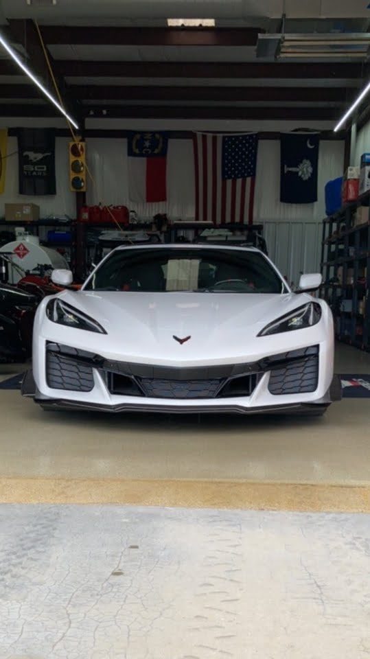 A white sports car is parked in a garage with various flags hanging on the wall, including the American flag, under fluorescent lighting.
