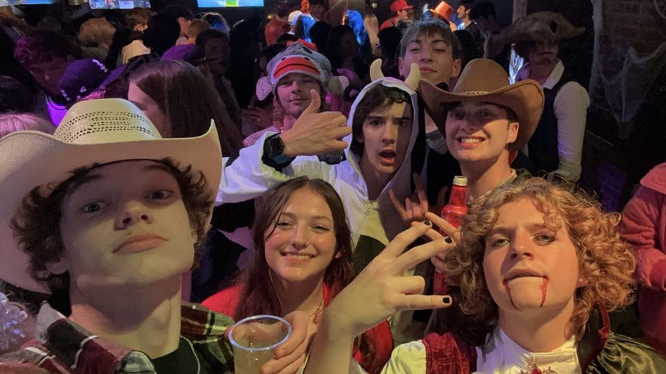 A group of young people in costumes enjoys a party, posing for a selfie, with cowboy hats, fake blood, and playful expressions. They're indoors with a crowd behind.