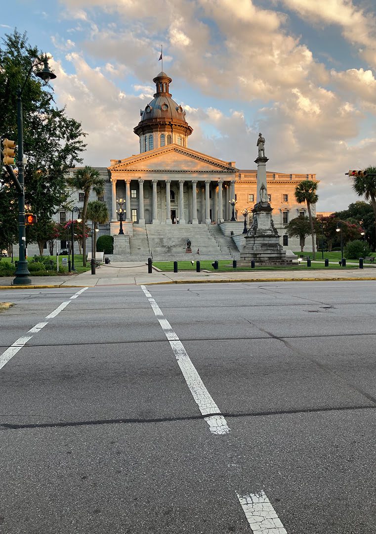 A domed building, possibly a capitol, with steps and statues at sunset. A pedestrian crossing and streetlamps frame the scene. Few people are visible.