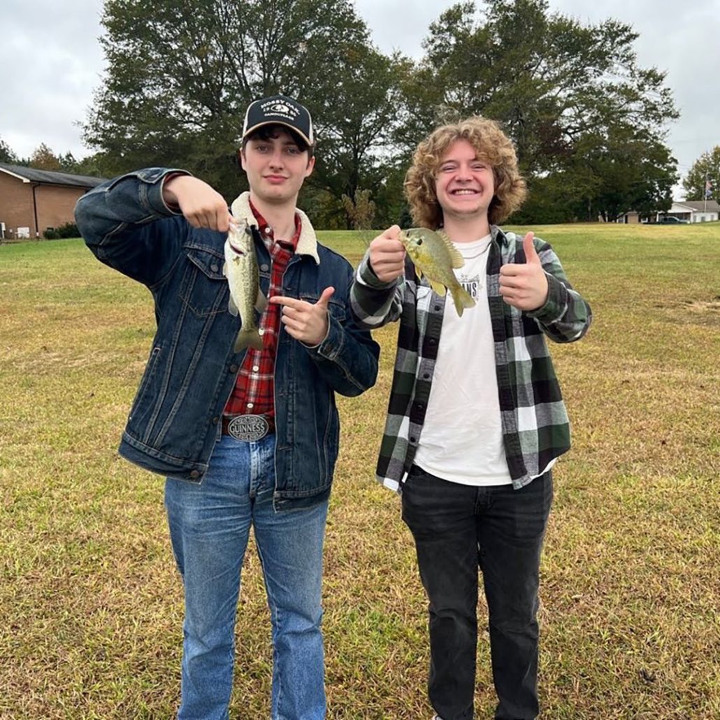 Two people stand outdoors holding fish they've caught. They're smiling, one giving a thumbs-up. Trees, grass, and a building are in the background.