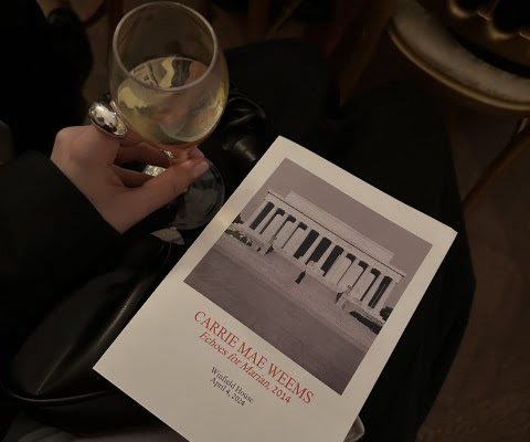 A person sitting with a glass of wine and a program titled "Carrie Mae Weems" for an event hosted by the Guggenheim Museum in 2014.