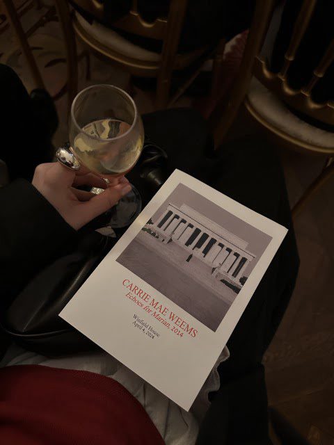 A person sitting with a glass of wine and a program titled "Carrie Mae Weems" for an event hosted by the Guggenheim Museum in 2014.