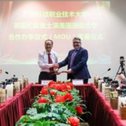 Two people are shaking hands at a formal event with red flowers on the table, Chinese text on a screen behind them, and an audience clapping.