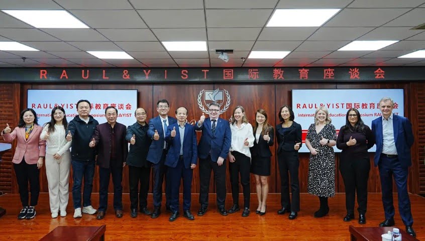 A group of people are standing in front of a backdrop with logos, giving thumbs up in a conference room, suggesting a collaborative or celebratory occasion.