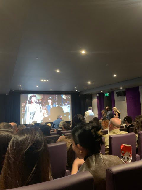 This is an indoor setting where people are seated in a cinema with purple chairs, watching a film on a large screen. Some individuals are standing at the back.