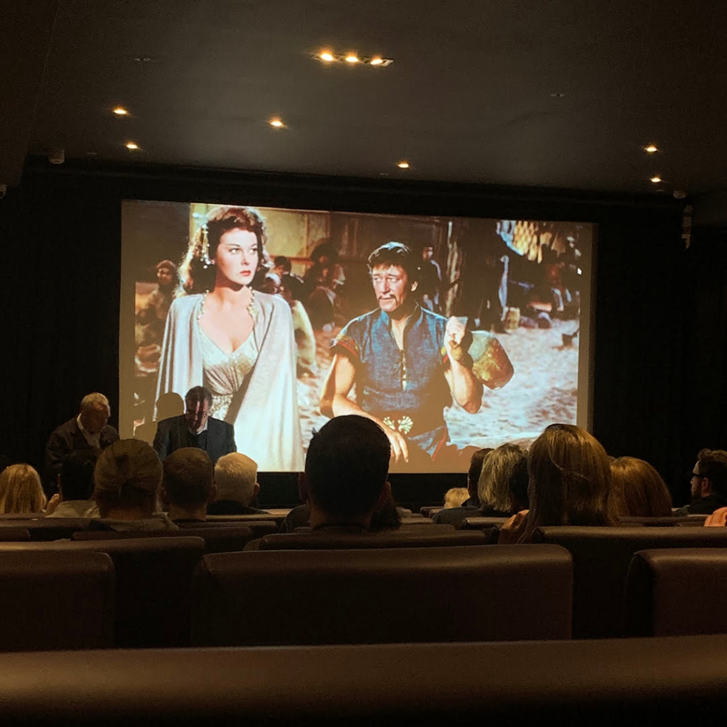 Audience in a dark cinema watching a classic film, projected on the screen, featuring two characters in historical costumes, one holding a drinking vessel.