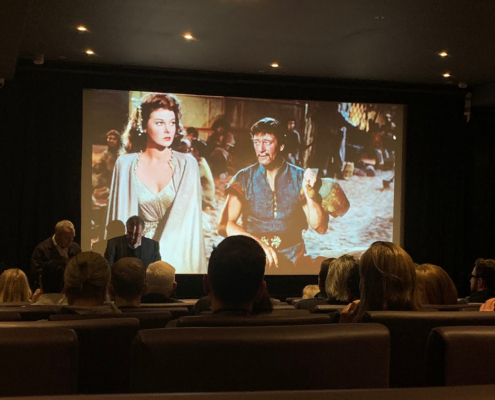 Audience in a dark cinema watching a classic film, projected on the screen, featuring two characters in historical costumes, one holding a drinking vessel.