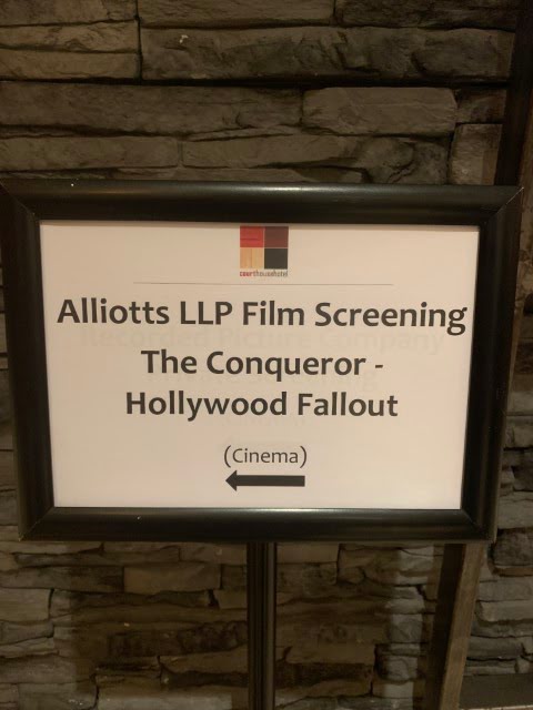 A framed sign for "Alliotts LLP Film Screening" of "The Conqueror - Hollywood Fallout" with an arrow indicating the cinema's direction, displayed against a stone wall background.
