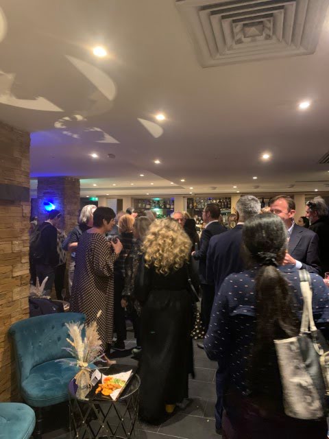 The image shows a group of people gathered at an indoor event, possibly a party or networking event, with dim lighting and a cozy atmosphere.