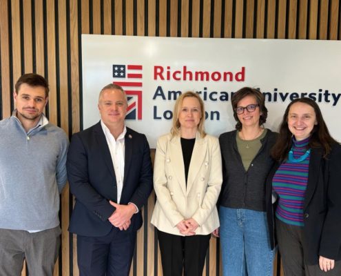 Five people are standing in front of a sign reading "Richmond American University London," smiling casually for a photo in a wood-paneled room.