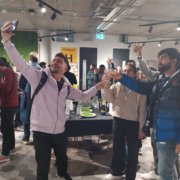 In an indoor space, people are standing around a table with drinks, raising their glasses. One person is taking a selfie, and others are cheering.