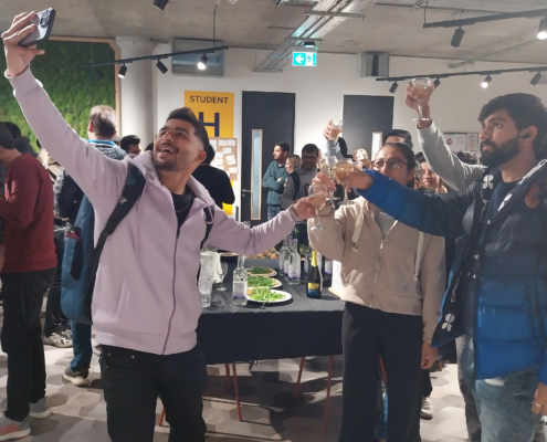 In an indoor space, people are standing around a table with drinks, raising their glasses. One person is taking a selfie, and others are cheering.