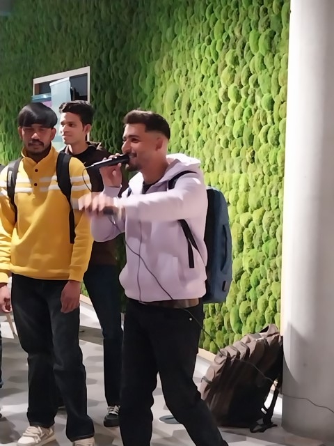 A person is singing into a microphone enthusiastically indoors. Two others watch from behind. A green textured wall and column are visible in the background.