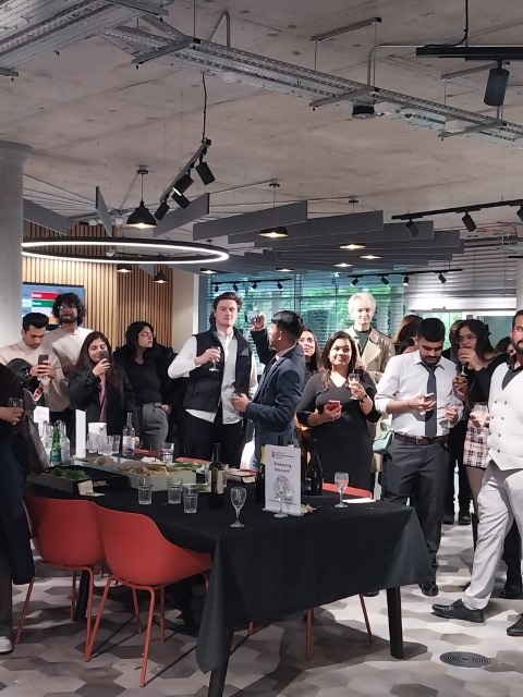 Group of people gathered in a modern interior with exposed ceiling, engaged in conversation, holding drinks, with a bar table in the foreground.
