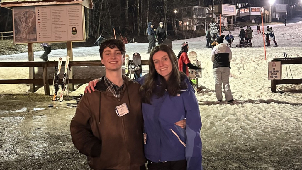 Two people are smiling in front of a snowy ski slope with skiers and snowboarders at night; ski equipment is visible to the left.