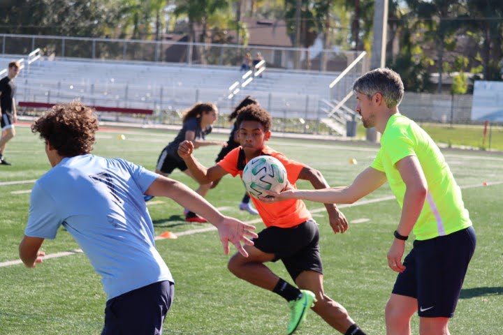 Three individuals are playing soccer on a sunny day with stadium bleachers in the background. One person is controlling the ball closely.