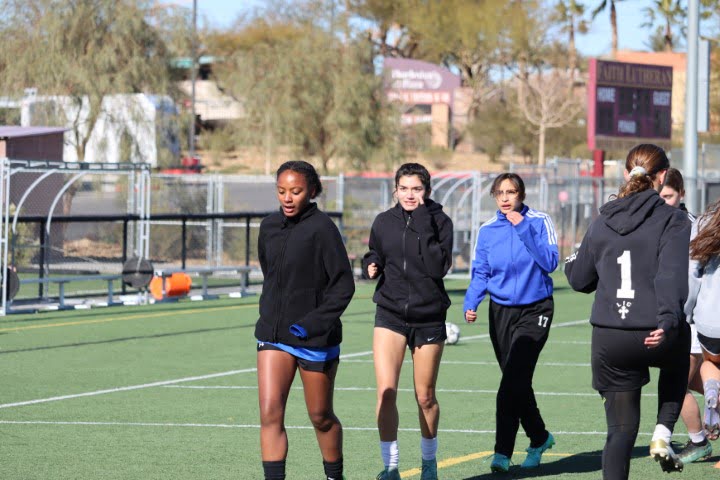 A group of people on a sports field, some in athletic wear, potentially a team engaged in pre-game or practice exercises, with surrounding sports facilities.