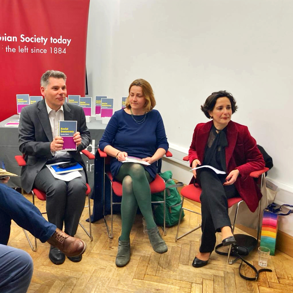Three people sit in front of a red banner reading "Fabian Society today" holding books. They're engaged in a discussion or presentation in a casual setting.