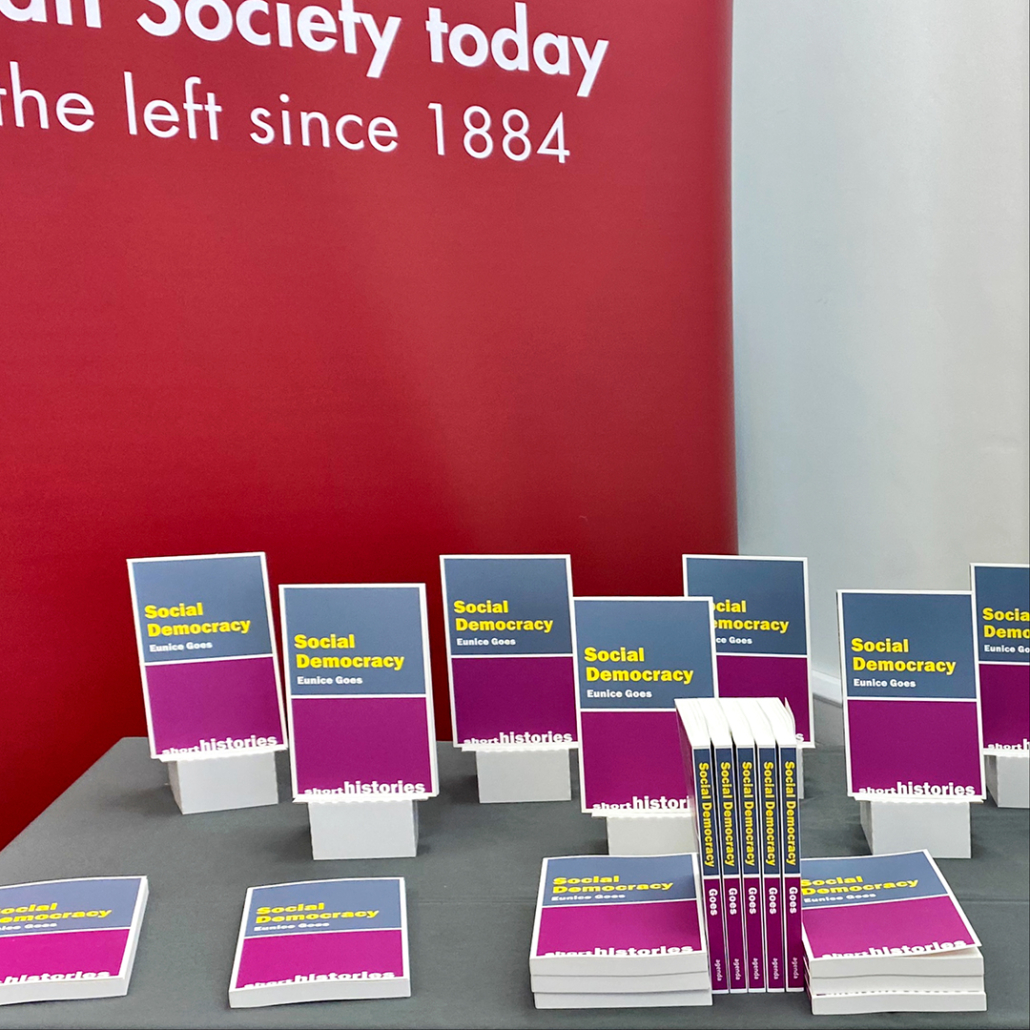 A display of books titled "Social Democracy" by Eunice Goes, in front of a red backdrop with text "Join Society today - the left since 1884."