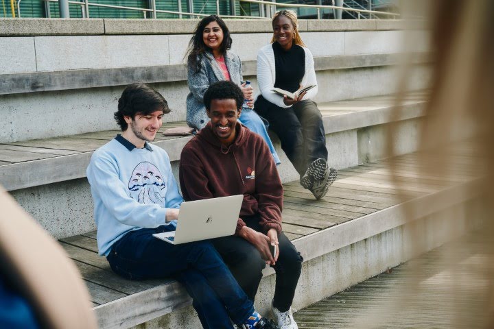 Four people are sitting on outdoor steps. Two are looking at a laptop, smiling. The other two are behind them, enjoying a sunny day.
