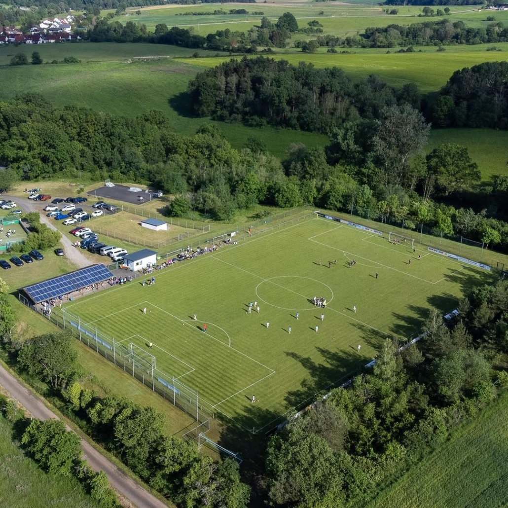 Aerial view of a soccer game in progress on a green field, with spectators, parked cars, and surrounding trees in a rural area.