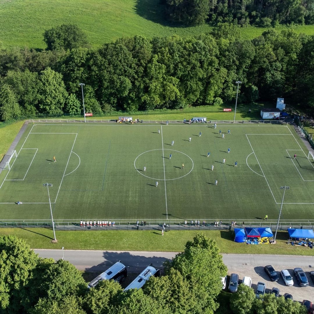 Aerial view of a soccer match in progress on a synthetic field, surrounded by trees with spectators and parked cars nearby.