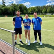 Three people are standing on a soccer field, dressed in blue sportswear, smiling, with one holding a small trophy and a bundle of colorful items.