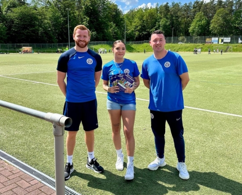 Three people are standing on a soccer field, dressed in blue sportswear, smiling, with one holding a small trophy and a bundle of colorful items.