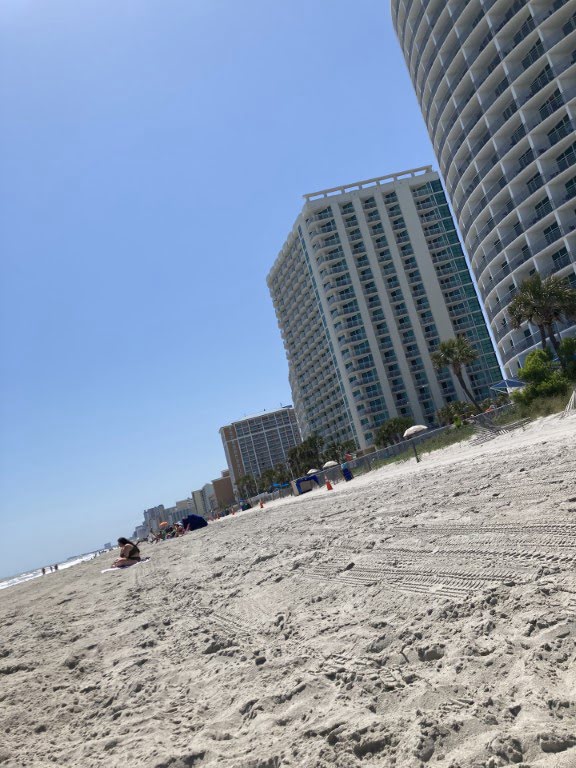 A sunny beach scene with tall buildings lining the shore, clear blue sky above, and a few people and beach equipment scattered on the sand.
