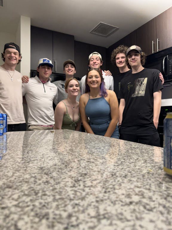 Seven young people pose smiling in a modern kitchen. Countertop in the foreground, wooden cabinets, and a microwave visible in the background.