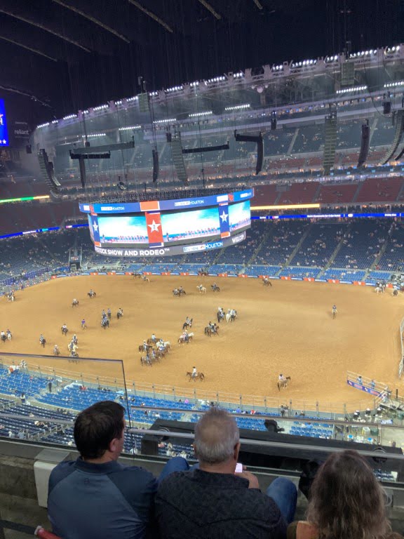 This image shows an indoor rodeo arena with several people on horses, large screens, spectators in the stands, and a dirt-covered ground.
