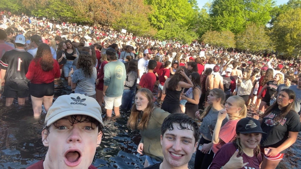 A dense crowd of young adults is gathered in a sunny outdoor setting, many standing in water, with cheerful expressions and casual attire.