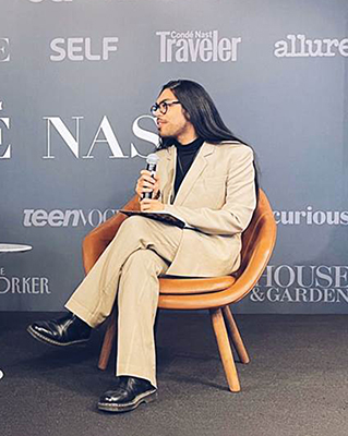 A person with long hair, wearing glasses, seated on a wooden chair, dressed in a beige suit, holding a microphone, against a branded backdrop.