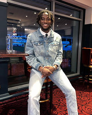 A person with long dreadlocks is smiling, standing indoors wearing a denim jacket, white pants, and a patterned shirt. A bar area with stools is visible behind.