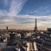 A scenic view of Paris, with the Eiffel Tower prominent among the city's rooftops under a wide sky streaked with clouds.